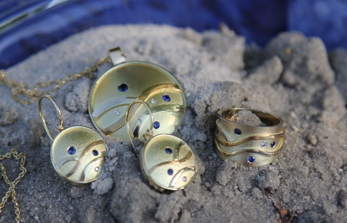 Set van gouden hanger, oorbellen en ring met golvende lijnen, blauwe steentjes en diamantjes, op zand